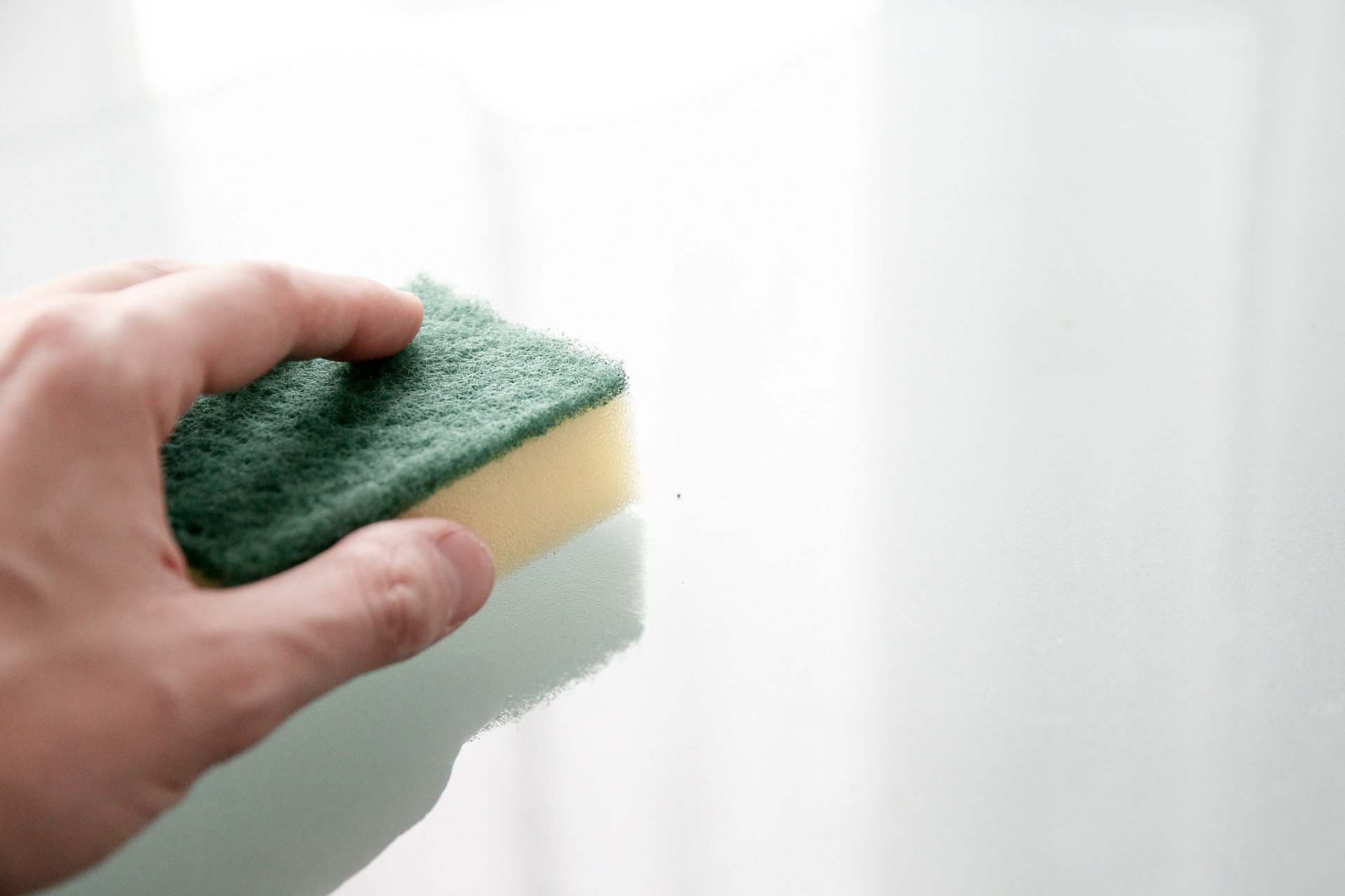 Hand wiping down a white counter with a green and yellow sponge.